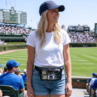 Woman at a baseball game wearing a white t-shirt and blue cap, with a crowd and stadium in the background.